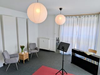 Heller Unterrichtsraum mit Notenständer, E-Piano mit aufgeschlagenen Noten, zwei Sesseln und Papierlampen