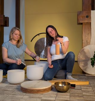 Zwei Frauen sitzen am Boden zwischen Klangschalen, Gong und Rahmentrommel; eine singt mit geschlossenen Augen und hält ein Koshi-Klangspiel