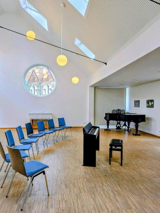Heller Saal mit hohem Dach, rundem Fenster und Kugellampen; blaue Stühle im Halbkreis vor Flügel und E-Piano